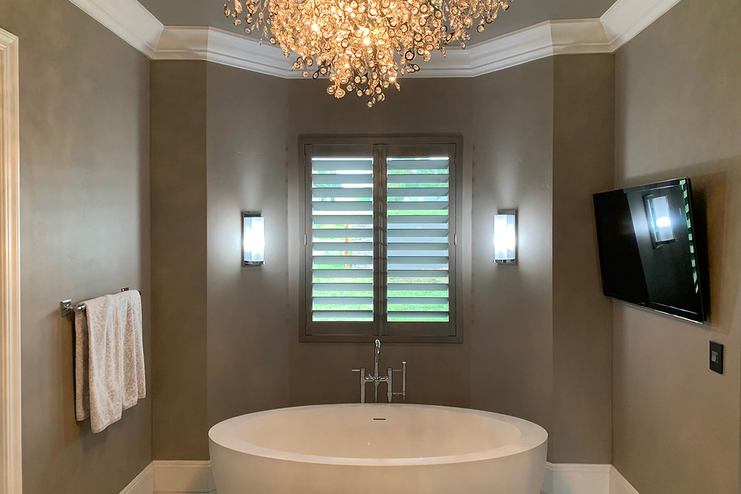 Elegant bathroom with a TV and chandelier, showing plantation shutters covering the window.