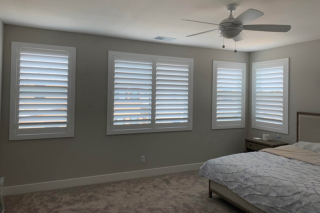 A large, white, simple bedroom with plantation shutters half open on the four windows.