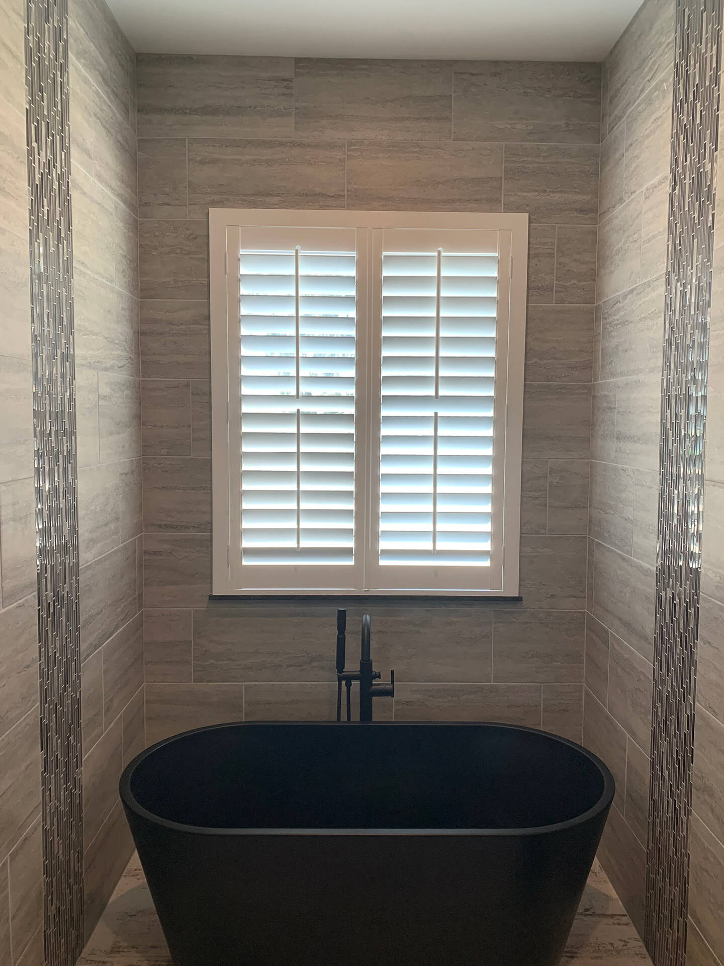 A black bathtub with a window above it covered by shutters.