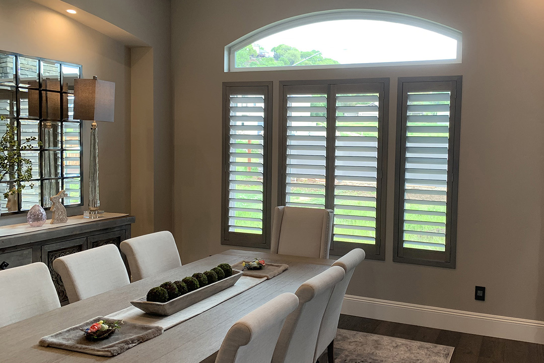 An elegant dining room with gray plantation shutters open to show the front lawn.